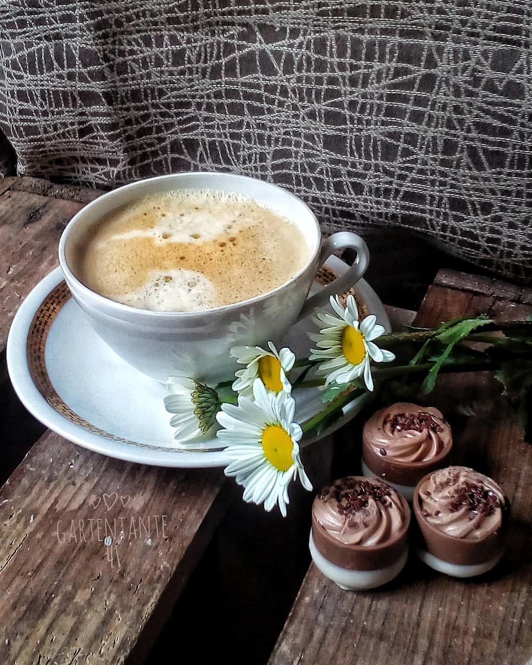Чашка кофе с пеной на блюдце, рядом лежат ромашки и три шоколадных конфеты на деревянной поверхности.